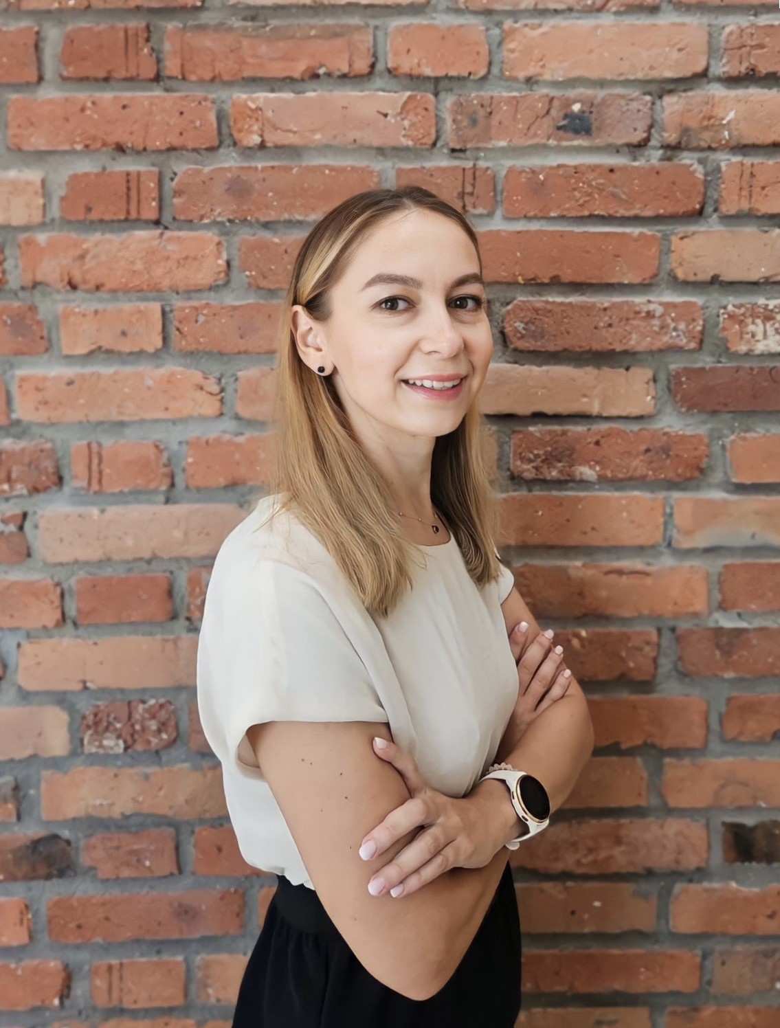 Portret uśmiechniętej kobiety z włosami w kolorze jasnego blondu, stojącej na tle ściany z czerwonej cegły. Kobieta ma skrzyżowane ramiona i opuszczone dłonie, z zegarkiem na lewym nadgarstku.