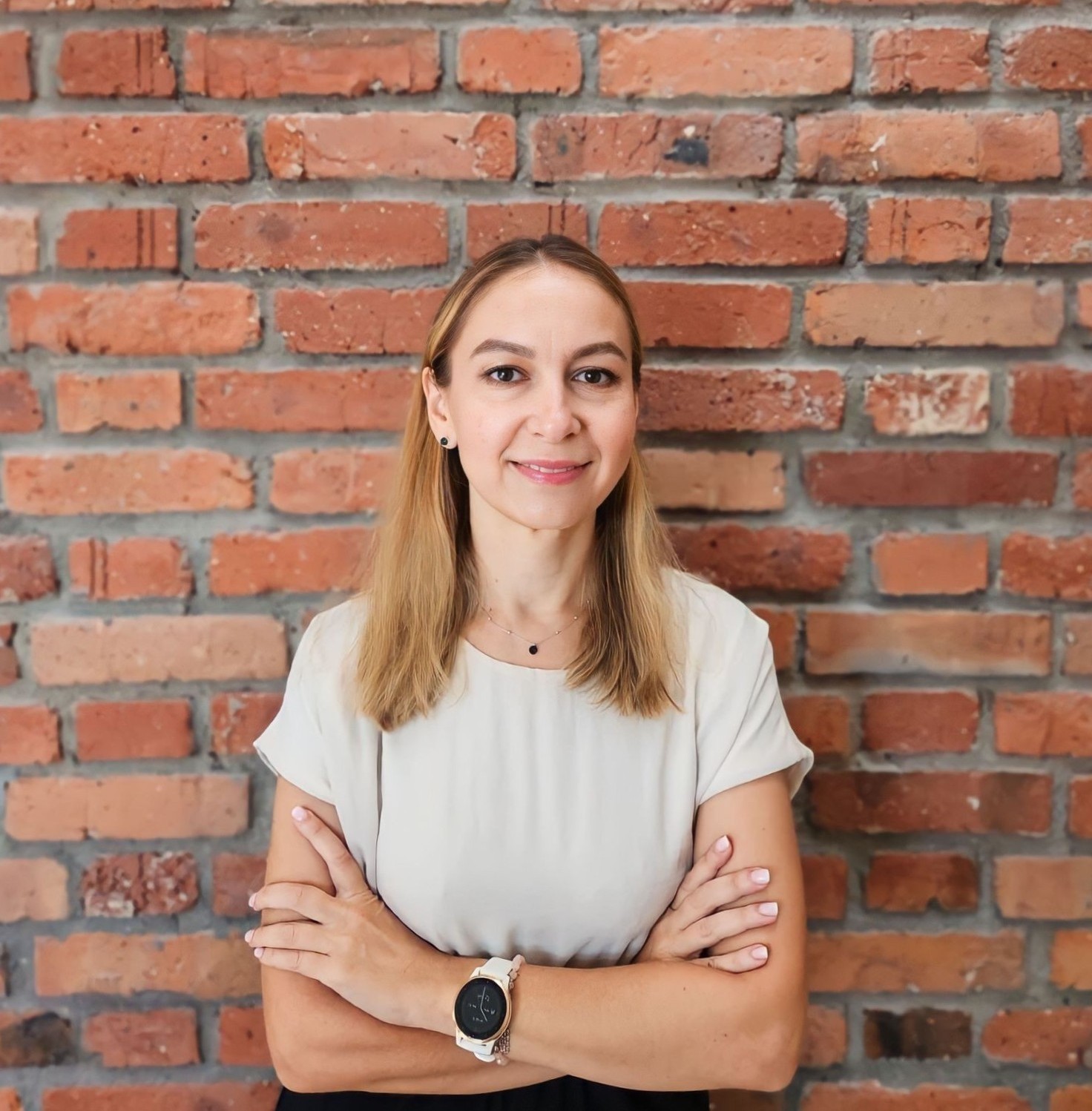Portret uśmiechniętej kobiety z włosami w kolorze jasnego blondu, stojącej na tle ściany z czerwonej cegły. Kobieta ma skrzyżowane ramiona na wysokości klatki piersiowej i nosi zegarek na lewym nadgarstku.